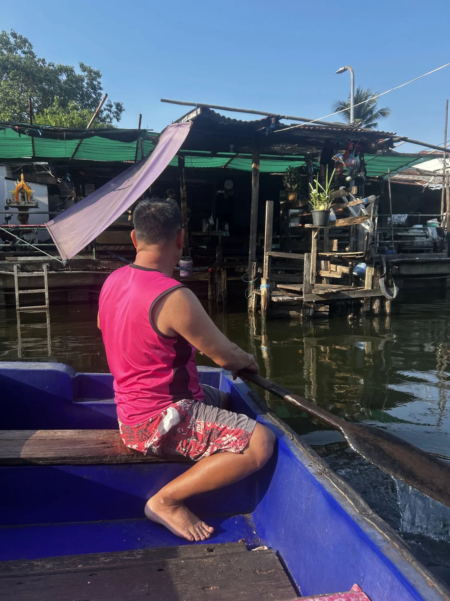 A boatman in a pink tank top paddles the author across a Bangkok river in a purple boat with a purple cloth sail, reading the current with a single wooden oar, floating wooden houses and a gold spirit shrine behind him