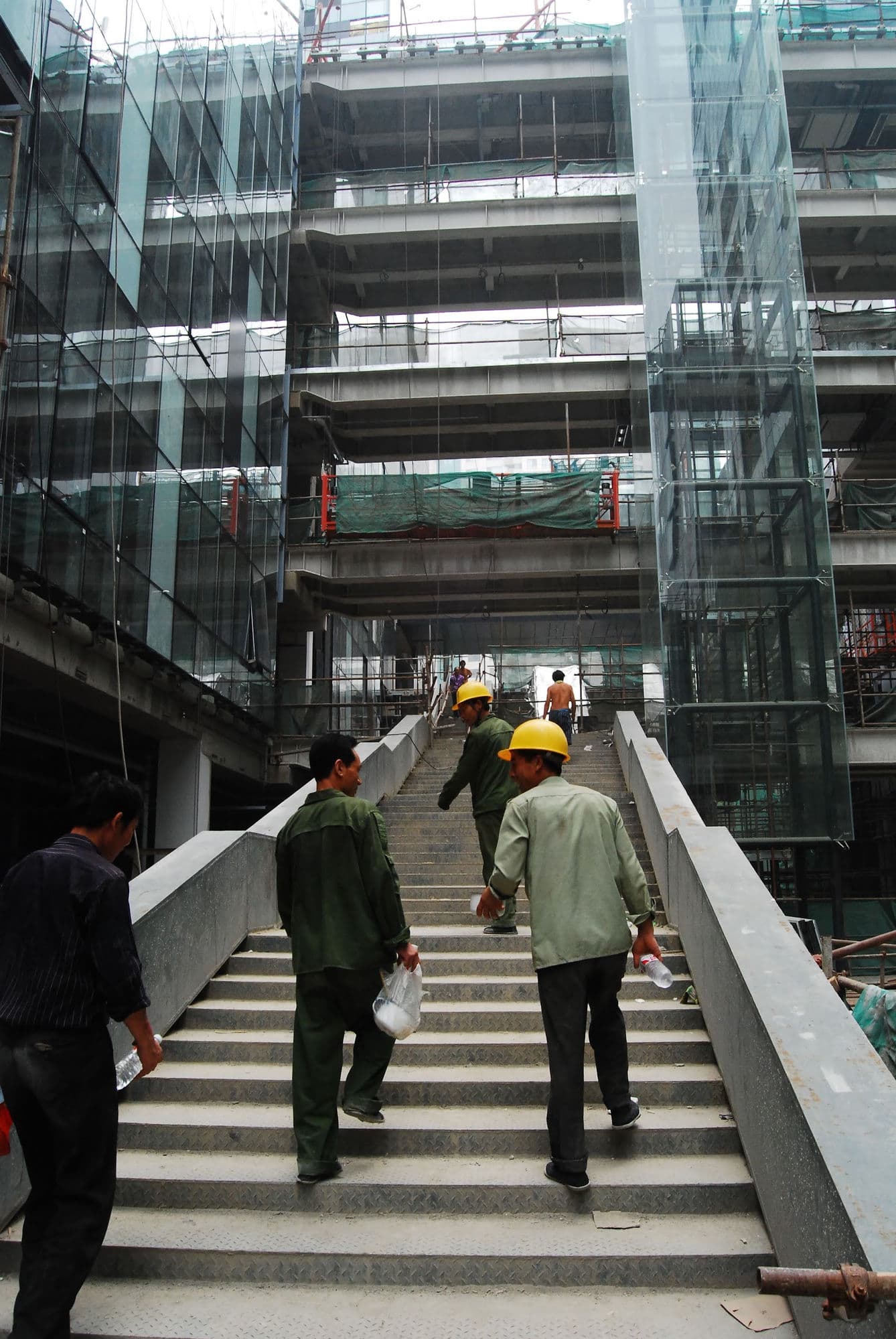 Construction workers in hard hats climbing a concrete staircase inside a glass tower under construction, China