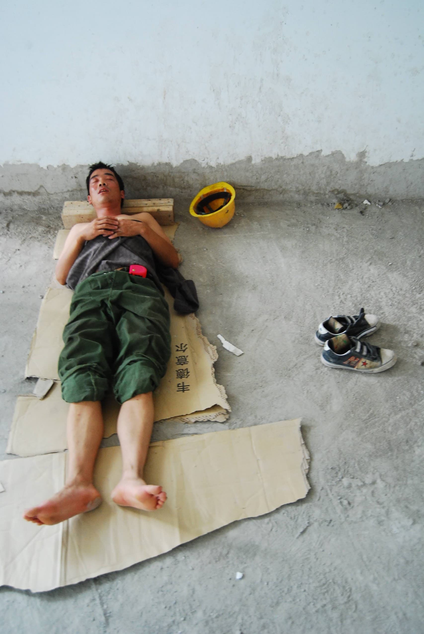 A construction worker asleep on flattened cardboard at a site, his hard hat and worn shoes beside him