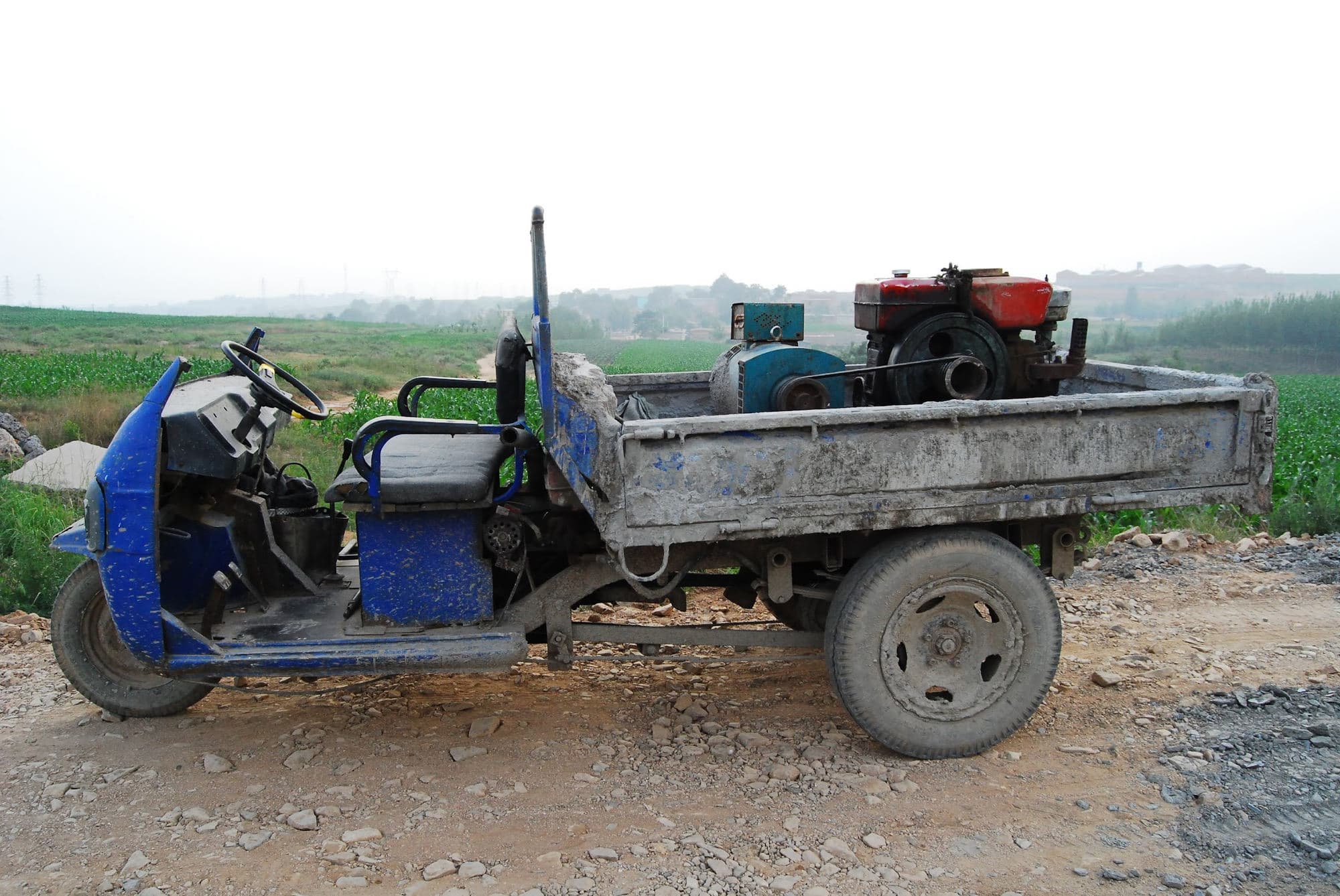A dusty blue three-wheeled diesel cargo truck parked on a gravel road by farm fields, rural China